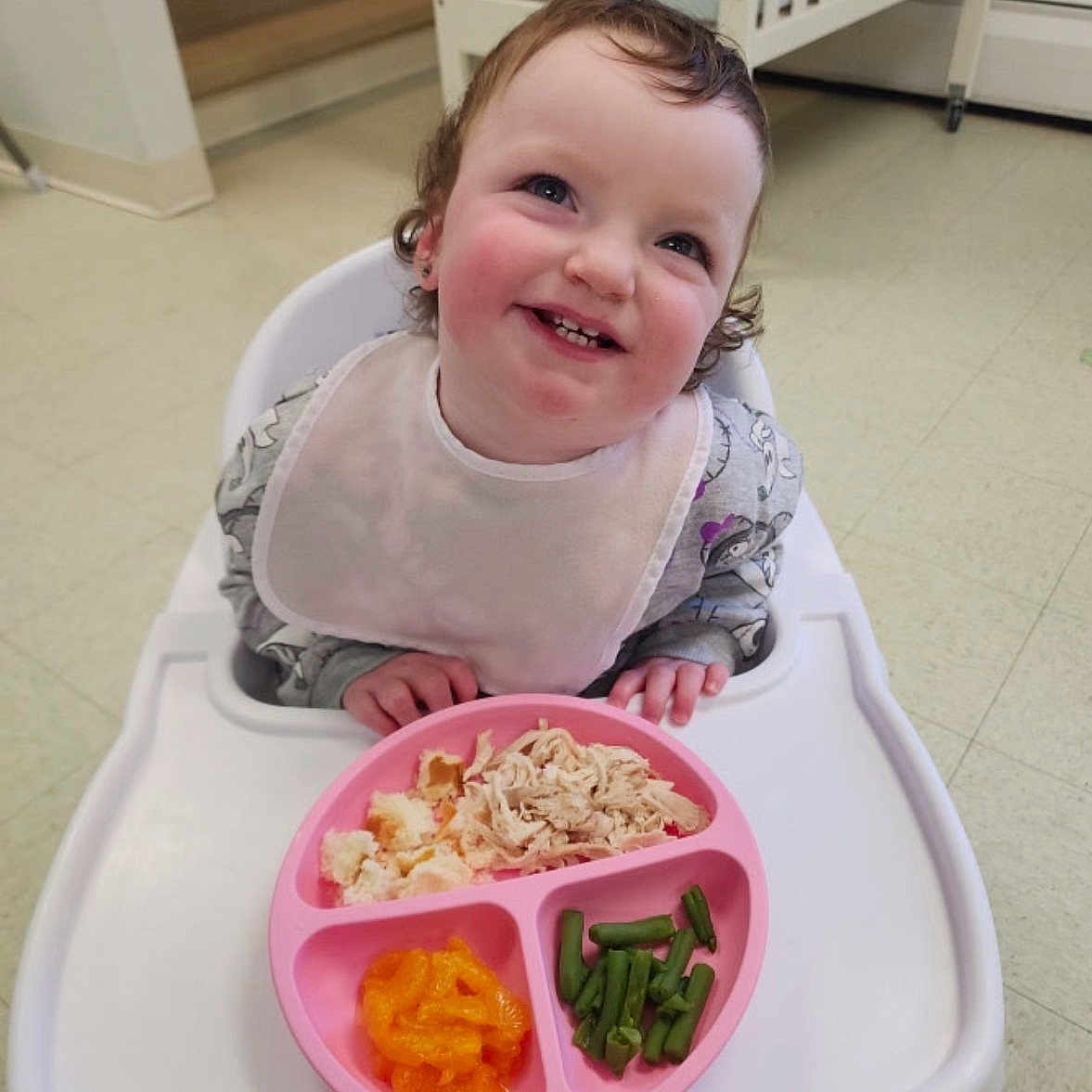 Navaya is registered to the contest to win money with this photo: bib, child, crib, curly_hair, eating, floor, food, green_beans, happy, high_chair, indoor, mandarin_oranges, meal, person, pink_plate, plate, shredded_chicken, smiling, stairs, toddler