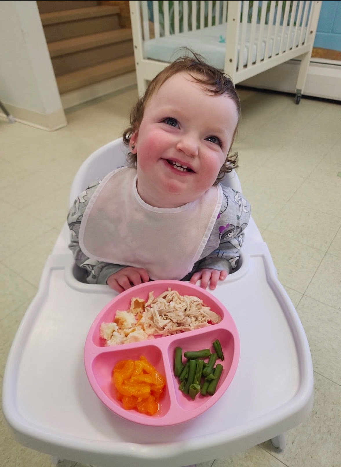 Navaya is registered to the contest to win money with this photo: toddler, child, high_chair, bib, food, plate, shredded_chicken, green_beans, mandarin_oranges, smiling, indoor, crib, stairs, floor, happy, eating, curly_hair, person, meal, pink_plate
