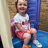 brick_wall, casual_clothing, child, colorful, curly_hair, cute, footwear, happy, outdoor, person, plastic_play_structure, play, playground, red_shorts, shoes, sitting, smiling, toddler, white_shirt, young_child