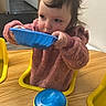 toddler, child, indoor, table, sippy_cup, blue_bowl, pink_sweater, yellow_chair, wooden_table, face, hair, person, eating, drinking, baby, room, wall_art, playroom, furniture, curly_hair