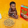 baby, toddler, child, smiling, laughing, cereal, bowl, milk, glass_bottle, food, breakfast, yellow_background, cereal_box, reese's_puffs, happy, sitting, indoors, cute, playful, person