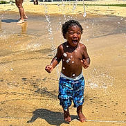 Zayyvon joined the competition — help win amazing prizes! toddler, child, water, splash_pad, playing, outdoor, summer, fun, joyful, splashing, water_droplets, barefoot, blue_shorts, park, sunny, happy, person, curly_hair, recreation, family