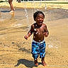 toddler, child, water, splash_pad, playing, outdoor, summer, fun, joyful, splashing, water_droplets, barefoot, blue_shorts, park, sunny, happy, person, curly_hair, recreation, family