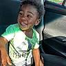 child, toddler, smiling, car_interior, seat, window, sunlight, green_shirt, shorts, happy, face, person, seatbelt, hair, skin, daylight, vehicle, casual_clothing, indoor, portrait