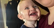 Marlin is registered to the contest to win money with this photo: baby, child, person, smile, glasses, indoor, happy, holding, shirt, face, blue_eyes, short_hair, clothing, coats, background, closeup, cute, joyful, family, portrait