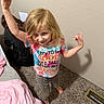 child, girl, smiling, tie_dye_shirt, colorful_clothing, indoor, carpet, blanket, person, snack, barefoot, wall, vent, cheeky, happy, playful, long_hair, casual_clothing, holding_object, room