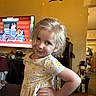 child, girl, smile, pose, floral_dress, indoor, television, cartoon, living_room, furniture, wood_floor, cabinet, toy, person, hair, hand_on_hip, cute, playful, happy, home