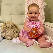Carsen is registered to the contest to win money with this photo: baby, child, infant, pink_dress, striped_clothing, plush_toy, stuffed_animal, bunny, cushioned_chair, knitted_blanket, beige_chair, smiling, sitting, cute, portrait, indoor, person, holding_sign, 8_months, happy