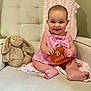 baby, child, infant, pink_dress, striped_clothing, plush_toy, stuffed_animal, bunny, cushioned_chair, knitted_blanket, beige_chair, smiling, sitting, cute, portrait, indoor, person, holding_sign, 8_months, happy