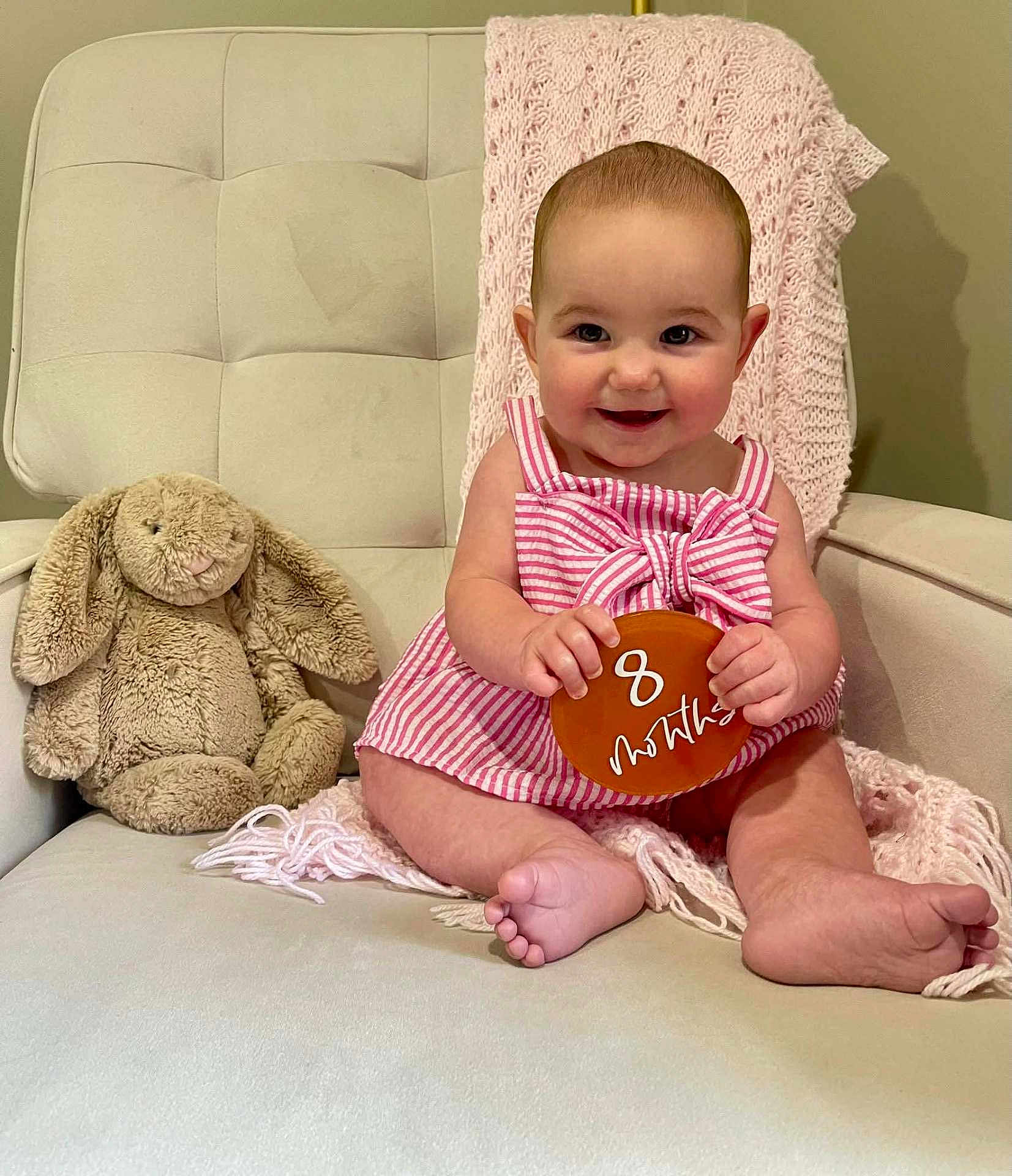 Carsen is registered to the contest to win money with this photo: baby, child, infant, pink_dress, striped_clothing, plush_toy, stuffed_animal, bunny, cushioned_chair, knitted_blanket, beige_chair, smiling, sitting, cute, portrait, indoor, person, holding_sign, 8_months, happy