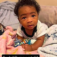 Cy'Meir'amor is registered to the contest to win money with this photo: baby, child, infant, portrait, sitting, lying_down, curious, face, eyes, hair, smiling, indoors, bed, blanket, plush_toy, clothing, bottle, home, photograph, text_overlay
