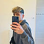 selfie, mirror, young_person, curly_hair, hoodie, bag, bathroom, radiator, bathtub, towel, phone, hand, casual_clothing, indoor, portrait, reflection, tile, wall, fashion, youth