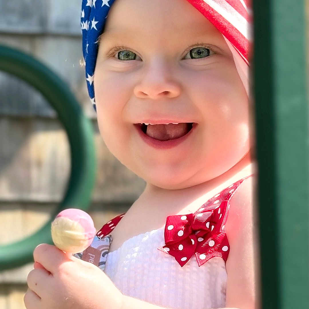 Naveigh is registered to the contest to win money with this photo: american_flag, baby, child, cute, face, hand, happy, headband, lollipop, outdoor, play, playground, polka_dot, portrait, red_dress, smiling, summer, sunlight, toddler, young_child