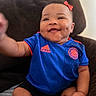 baby, child, infant, smiling, happy, face, portrait, soccer_jersey, blue_clothing, red_bow, sitting, couch, indoor, home, close_up, adorable, cheeks, dimples, photography, sportswear