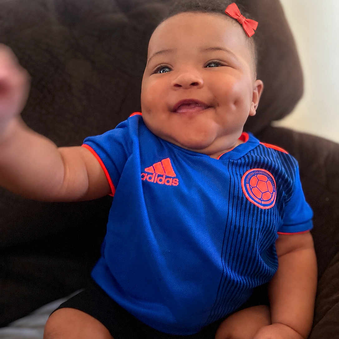 Juliana joined the competition — help win amazing prizes! baby, child, infant, smiling, happy, face, portrait, soccer_jersey, blue_clothing, red_bow, sitting, couch, indoor, home, close_up, adorable, cheeks, dimples, photography, sportswear