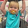 toddler, child, pacifier, shopping_cart, hat, blue_shirt, blond_hair, store, baby_clothes, smiling, happy, arms_up, indoor, retail, clothing_rack, cute, person, face, playful, young_child