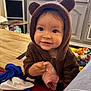 bear_ears, brown_hoodie, cabinet, child, clothing, cozy, curious, cute, face, furniture, happy, home, indoor, person, pink_toy, playtime, smiling, toddler, toy, wooden_table