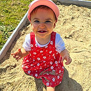 Aria a rejoint le concours — aidez-le/la à gagner de superbes lots ! child, toddler, baby, sandbox, sand, pink_hat, red_dress, floral_pattern, smile, teeth, outdoors, grass, sunlit, playtime, hands, bare_foot, bucket, curious, happy, portrait
