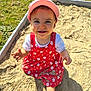 child, toddler, baby, sandbox, sand, pink_hat, red_dress, floral_pattern, smile, teeth, outdoors, grass, sunlit, playtime, hands, bare_foot, bucket, curious, happy, portrait