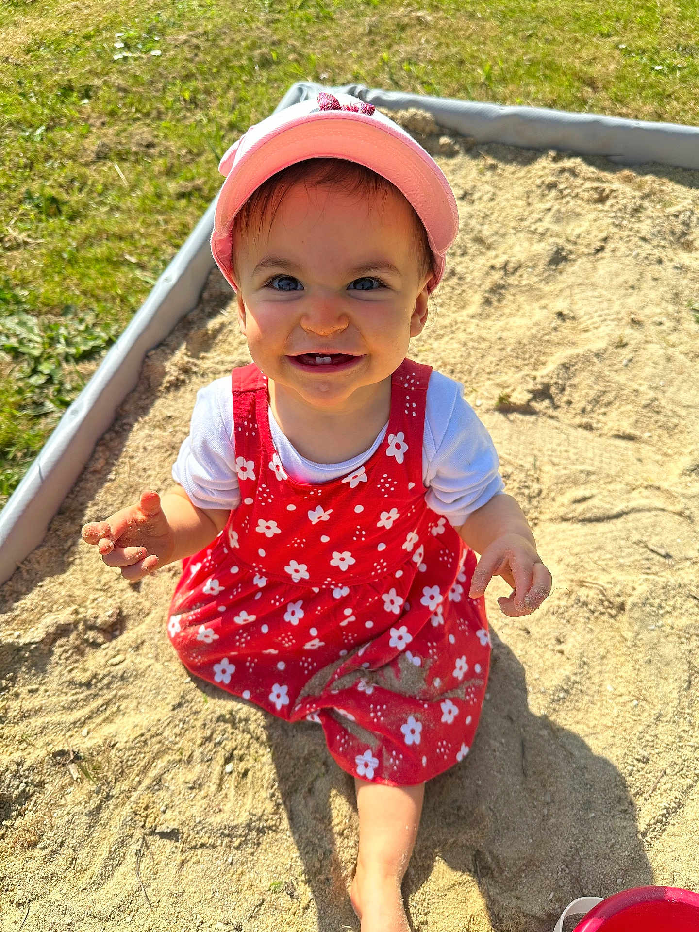 Aria a rejoint le concours — aidez-le/la à gagner de superbes lots ! child, toddler, baby, sandbox, sand, pink_hat, red_dress, floral_pattern, smile, teeth, outdoors, grass, sunlit, playtime, hands, bare_foot, bucket, curious, happy, portrait