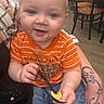 Zack is registered to the contest to win money with this photo: adult_arm, baby, blue_jeans, chairs, child, close_up, eating, family, food, happy, indoor, infant, orange_shirt, portrait, restaurant, seated, smiling, spoon, striped_clothing, tattoos