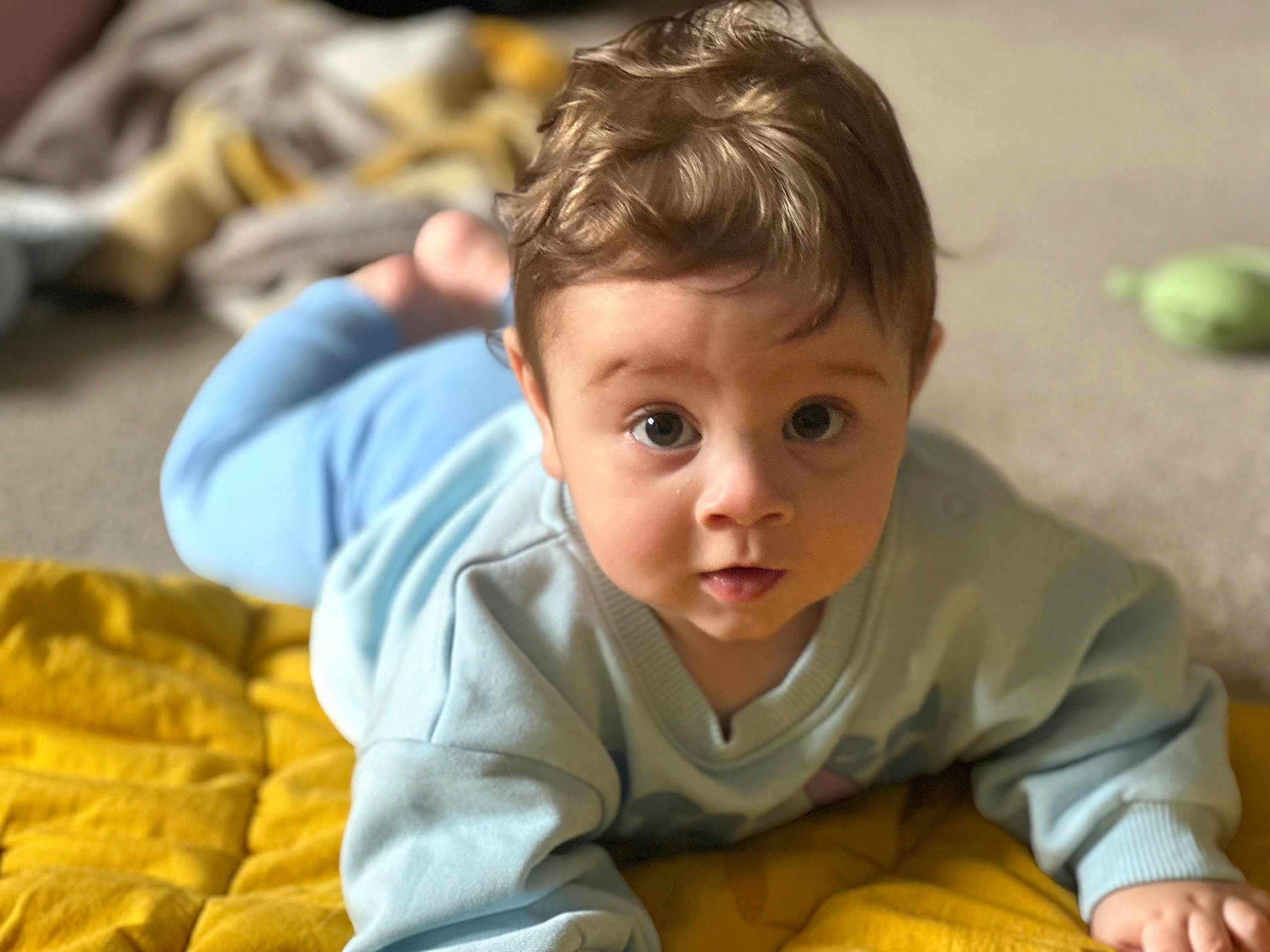 Cooper joined the competition — help win amazing prizes! baby, infant, child, blue_clothing, yellow_quilt, curly_hair, face, eyes, indoors, soft_texture, blanket, cute, portrait, lying_down, carpet, young_child, expression, crawling, toddler, cozy
