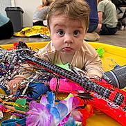 Cooper is registered to the contest to win money with this photo: baby, child, face, wide_eyes, pout, toy_guitar, party_props, streamers, yellow_surface, indoor, people_in_background, stroller, colorful, playful, curly_hair, pale_skin, focus_on_baby, blurred_background, floor, casual_clothing