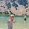 blurred_background, casual_clothing, child, curly_hair, fun, lake, mountain, nature, outdoor, person, playing, recreation, scenic, striped_clothing, summer, sunlight, wading, water, waterfront, young_child