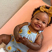 Bailey is registered to the contest to win money with this photo: baby, smiling, child, romper, sunflower, headband, bow, curly_hair, peach_cushion, happy, infant, cute, indoor, skin, hand, face, person, clothing, lying_down, adorable