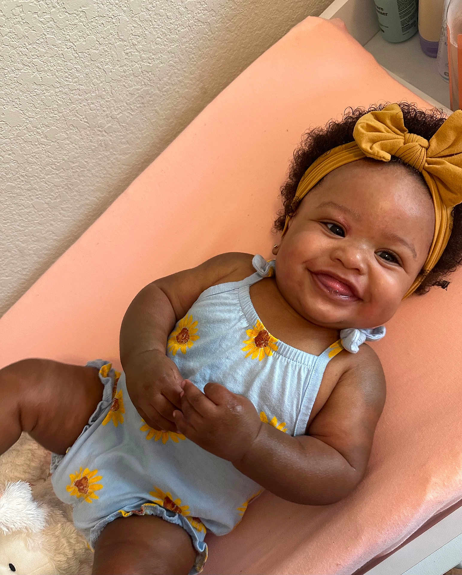 Bailey is registered to the contest to win money with this photo: baby, smiling, child, romper, sunflower, headband, bow, curly_hair, peach_cushion, happy, infant, cute, indoor, skin, hand, face, person, clothing, lying_down, adorable