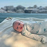 Salvateo is registered to the contest to win money with this photo: baby, infant, blanket, onesie, turtle_pattern, face, lying_down, outdoor, water, rocks, wooden_surface, soft_light, shadow, young_child, curious, portrait, cozy, cute, peaceful, nature