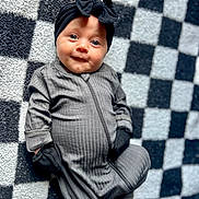 Layla joined the competition — help win amazing prizes! baby, infant, onesie, black_bow, headband, mittens, checkered_blanket, smile, face, blue_eyes, child, cute, lying_down, soft_texture, cozy, portrait, indoor, apparel, baby_clothes, adorable