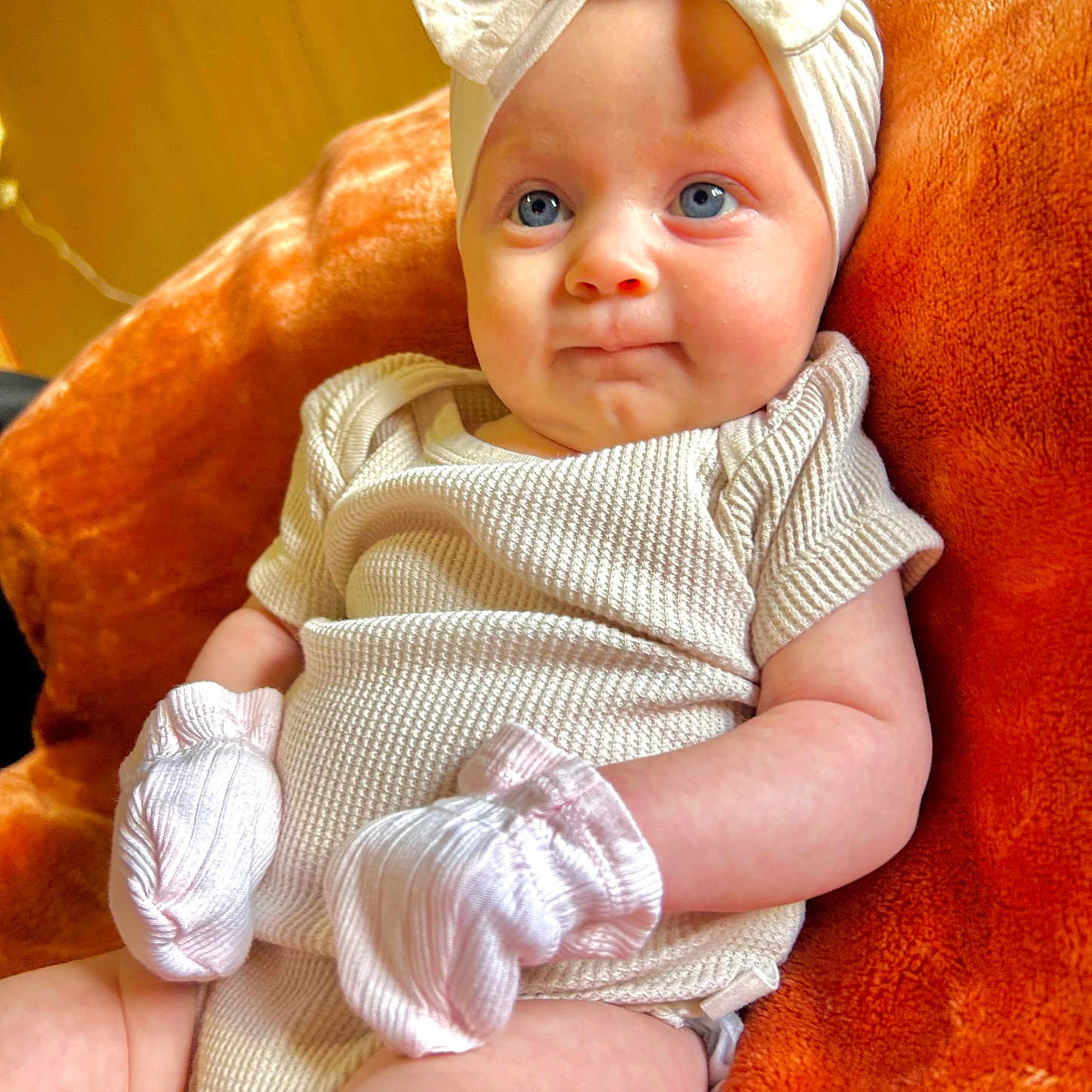 Layla joined the competition — help win amazing prizes! apparel, baby, blue_eyes, child, cozy, cream_outfit, cute, face, headband, indoors, infant, mittens, orange_blanket, portrait, sitting, skin, smile, soft_texture, white_bow, young