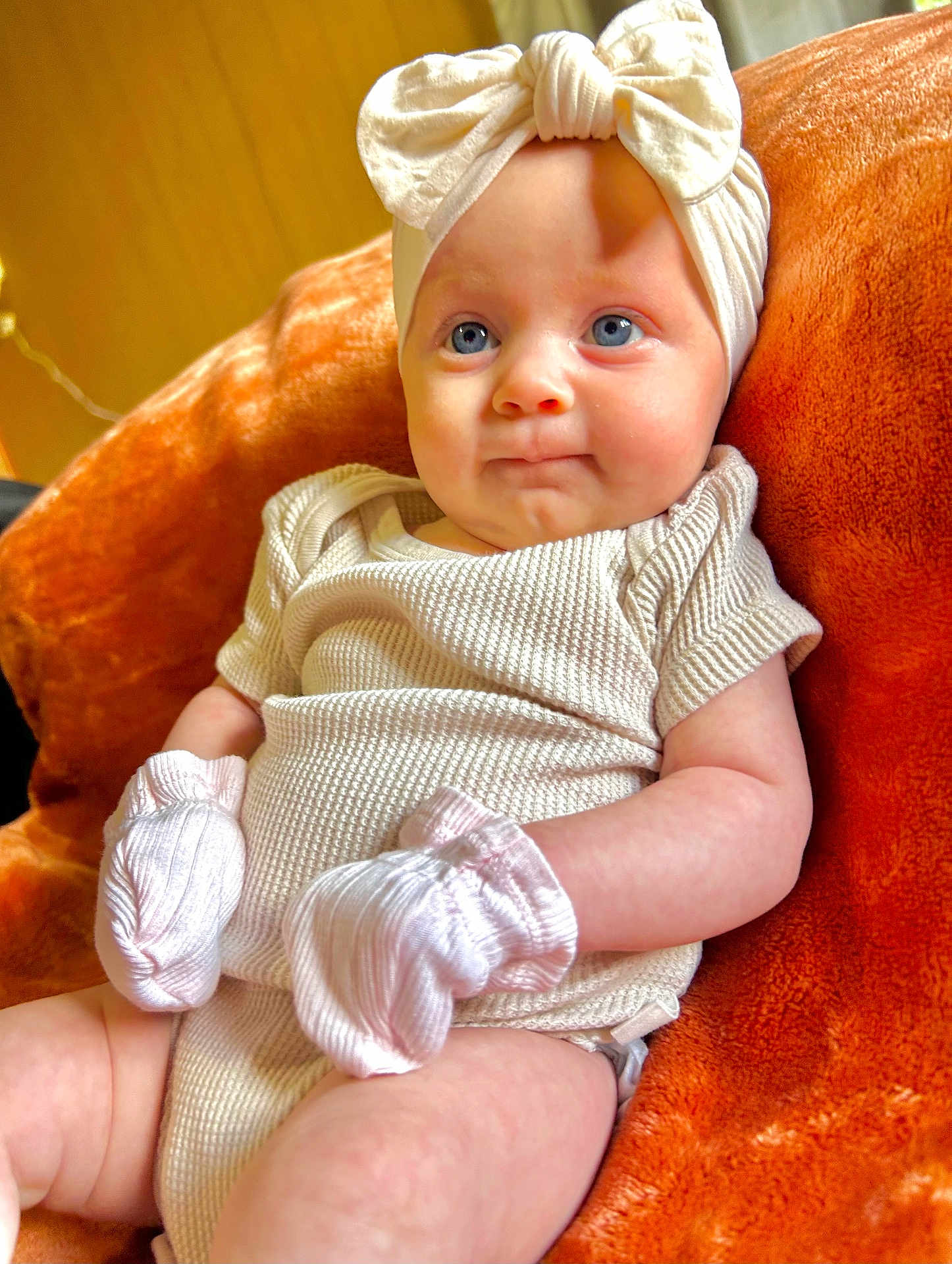 Layla joined the competition — help win amazing prizes! baby, infant, blue_eyes, white_bow, headband, mittens, cream_outfit, orange_blanket, soft_texture, cute, child, portrait, sitting, indoors, cozy, young, smile, skin, face, apparel