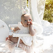 Aria joined the competition — help win amazing prizes! baby, child, crown, white_dress, chair, outdoor, sunlight, thumb_sucking, curtain, flowers, decor, portrait, cute, infant, soft_light, nature, seated, happy, celebration, half_birthday