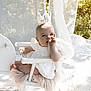 baby, child, crown, white_dress, chair, outdoor, sunlight, thumb_sucking, curtain, flowers, decor, portrait, cute, infant, soft_light, nature, seated, happy, celebration, half_birthday