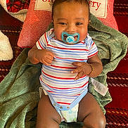 Amarion joined the competition — help win amazing prizes! baby, pacifier, striped_onesie, bracelet, green_blanket, red_cushion, carpet, smiling, infant, lying_down, cute, indoors, happy, child, soft_texture, cozy, portrait, person, toddler, clothing