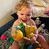 toddler, child, baby, curly_hair, smiling, stuffed_animals, toys, bottle, bed, blanket, indoor, happy, playful, person, cuddly_toys, colorful, cute, young_child, infant, room