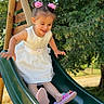 child, girl, slide, playground, outdoor, dress, white_dress, pink_flower, hair_clips, smiling, happy, summer, sunlight, green_slide, nature, trees, playing, fun, cute, footwear