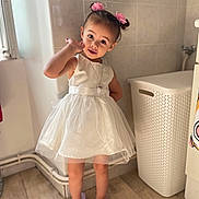 Lola participe au concours pour gagner de l'argent avec cette photo : child, girl, white_dress, pink_hair_accessories, slippers, indoor, floor, wooden_floor, laundry_basket, tile_wall, cute, posing, standing, portrait, young_child, floor_pipe, necklace, hand_gesture, natural_light, curious