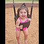 Angelina is registered to the contest to win money with this photo: toddler, child, swing, playground, smiling, happy, pink_clothing, wood_chips, outdoor, trees, grass, brick_wall, baby, person, play, fun, summer, daytime, park, hair_tie
