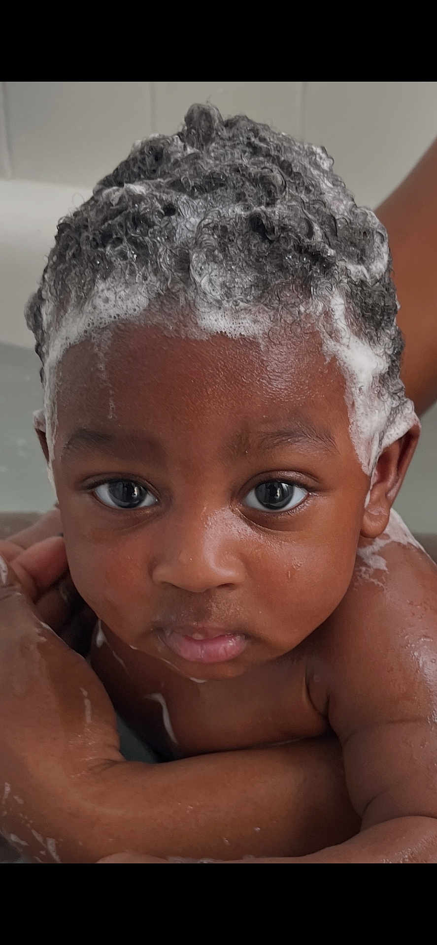 Arys is registered to the contest to win money with this photo: baby, child, bath, foam, shampoo, curly_hair, water, skin, face, eyes, expression, hand, adult_hand, wet, closeup, portrait, cute, indoors, personal_care, infant