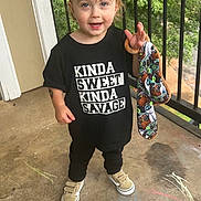 Grayson joined the competition — help win amazing prizes! balcony, black_tshirt, casual_clothing, chalk, child, concrete_floor, daylight, greenery, happy, outdoor, playful, railing, short_hair, slogan, smiling, sneakers, standing, toddler, toy, wooden_ring