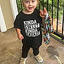 balcony, black_tshirt, casual_clothing, chalk, child, concrete_floor, daylight, greenery, happy, outdoor, playful, railing, short_hair, slogan, smiling, sneakers, standing, toddler, toy, wooden_ring