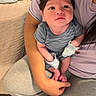 Jett is registered to the contest to win money with this photo: baby, infant, person, arm, clothing, striped_onesie, mittens, bed, blanket, adult, partial_face, indoor, home, comfort, cute, holding, light, skin, hair, sitting