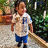Joandry is registered to the contest to win money with this photo: child, toddler, braided_hair, white_shirt, blue_shorts, sneakers, smiling, walking, plant, flowerpot, metal_fence, decorative_elements, colorful_lights, floor, indoor, happy, person, portrait, fashion, accessories