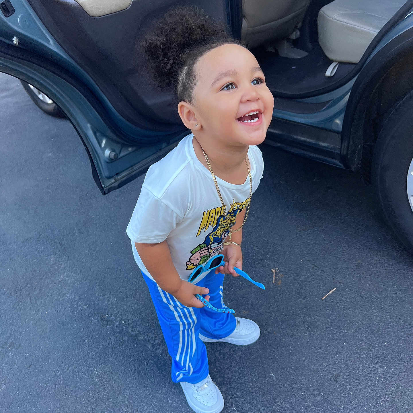 Joandry is registered to the contest to win money with this photo: toddler, child, smiling, happy, curly_hair, sunglasses, blue_pants, white_shirt, car, car_door, outdoor, asphalt, standing, casual_clothing, gold_chain, white_shoes, person, cute, joyful, playful