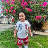 toddler, child, grass, flowers, bush, outdoor, playful, braided_hair, white_tshirt, red_shorts, sneakers, greenery, nature, portrait, cute, young_child, garden, smiling, standing, daylight