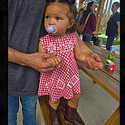 Catalina is registered to the contest to win money with this photo: child, toddler, pacifier, dress, boots, adult_hands, picnic_table, outdoor, park, trees, people, sunlight, bench, holding_hands, casual_clothing, hairstyle, daytime, red_and_white, cute, portrait
