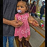 child, toddler, pacifier, dress, boots, adult_hands, picnic_table, outdoor, park, trees, people, sunlight, bench, holding_hands, casual_clothing, hairstyle, daytime, red_and_white, cute, portrait